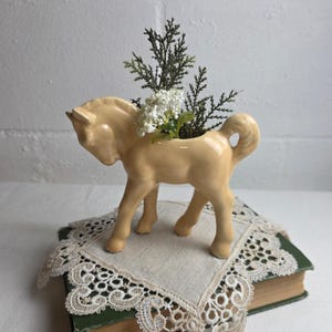 May include: A pale yellow ceramic horse-shaped planter with a glossy finish. The hollow back of the horse holds a small arrangement of green evergreen sprigs and tiny white flowers, creating a decorative piece for display.