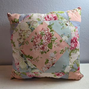 May include: A square throw pillow featuring a patchwork design with a central diamond panel. The fabric displays a floral pattern in shades of dusty pink, light blue, cream, and green. The construction consists of geometric fabric segments.