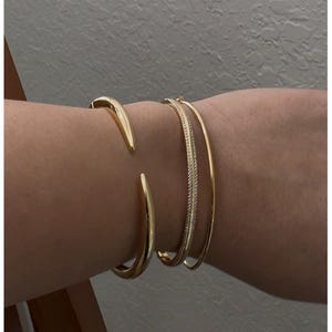 May include: A collection of gold-toned bracelets. The bracelets include a thick, open cuff, a thin, textured band, and a simple, smooth band. The jewelry is displayed on a person's arm, showcasing the different styles and designs.