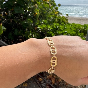 Peut inclure: Bracelet doré composé d'une série de formes ovales et rectangulaires reliées. Le bracelet est porté sur un poignet, avec en arrière-plan un feuillage vert, une plage et l'océan.