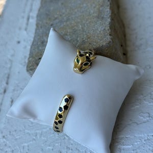 Può includere: Bracciale a polsino dorato con testa di giaguaro con occhi verdi. Il bracciale presenta un motivo geometrico nero e oro sulla fascia. Il gioiello è esposto su un cuscino bianco.