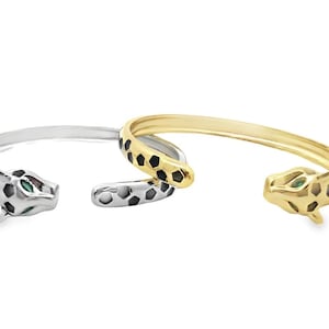 May include: Two open-ended cuff bracelets, one silver and one gold, each featuring a leopard head design. The leopard heads have black spots and green eyes. The bracelets are shown on a white background.