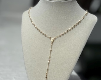 Peut inclure: Un chapelet doré composé de petites perles métalliques uniformes le long de la chaîne, d'une médaille centrale de forme ovale représentant une figure, et d'une petite croix simple suspendue au bas de la section verticale.