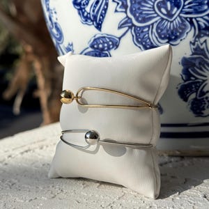 May include: Two minimalist bracelets, one gold-toned and one silver-toned, each featuring a spherical accent. They rest on a white, square pillow, set against a backdrop of a blue and white floral patterned ceramic vase.