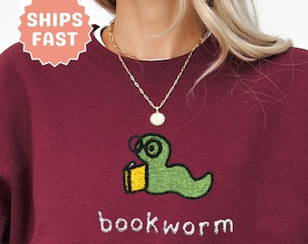 Puede incluir: Sudadera granate con un diseño de gusano de libro verde con gafas y un libro amarillo. La palabra "bookworm" está bordada en blanco. La sudadera tiene cuello redondo y mangas largas.