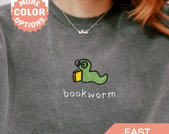 May include: A gray sweatshirt with an embroidered green bookworm design, wearing glasses and holding a book. The word "bookworm" is embroidered below the design. The image also includes text that says "Embroidered MORE COLOR OPTIONS" and "FAST SHIPPING".
