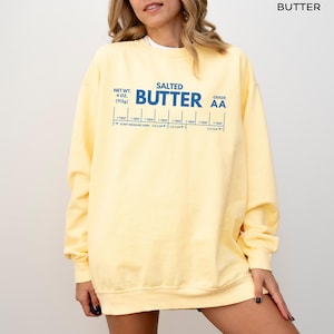 Op de afbeelding: Een lichtgeel sweatshirt met de tekst "SALTED BUTTER" in blauw. Het sweatshirt heeft ook een maattabel met de tekst "NET WT. 4 OZ (113g)" en "GRADE AA". Het sweatshirt is een casual stijl.