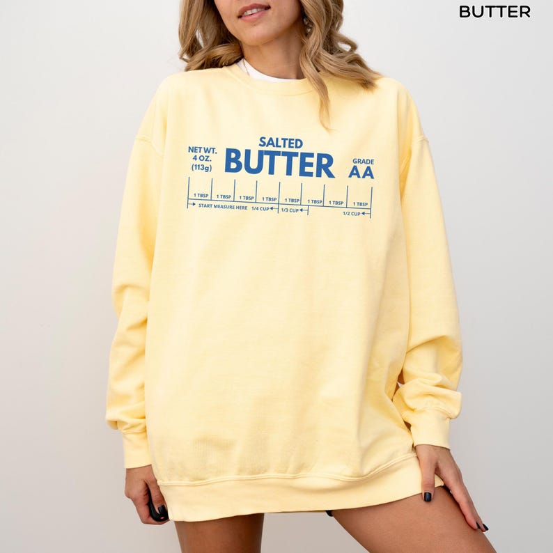 Op de afbeelding: Een lichtgeel sweatshirt met de tekst "SALTED BUTTER" in blauw. Het sweatshirt heeft ook een maattabel met de tekst "NET WT. 4 OZ (113g)" en "GRADE AA". Het sweatshirt is een casual stijl.