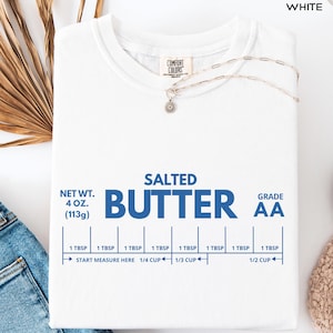 Op de afbeelding: Wit T-shirt met de blauwe woorden "SALTED BUTTER" en meetrichtlijnen. Het shirt heeft ook de tekst "NET WT. 4 OZ. (113g)" en "GRADE AA". Een zilveren ketting met een bloemhanger ligt op het shirt.