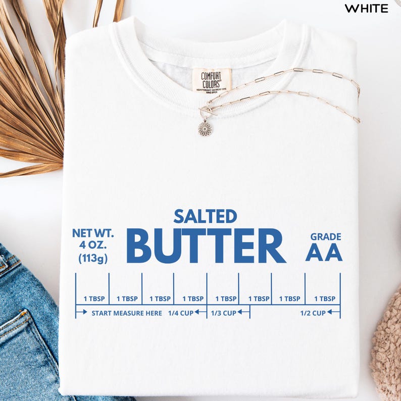 Op de afbeelding: Wit T-shirt met de blauwe woorden "SALTED BUTTER" en meetrichtlijnen. Het shirt heeft ook de tekst "NET WT. 4 OZ. (113g)" en "GRADE AA". Een zilveren ketting met een bloemhanger ligt op het shirt.