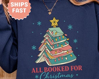 以下が含まれることがあります： 本で作られたクリスマスツリーのデザインが施されたネイビーブルーのスウェットシャツ。ライトと星で飾られています。「ALL BOOKED FOR *Christmas*」の文字が下にプリントされています。クルーネックと長袖です。