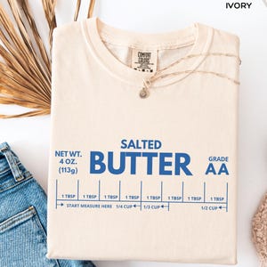 Op de afbeelding: Ivoor kleurig t-shirt met het woord "BUTTER" in grote blauwe letters. Het shirt heeft ook de tekst "SALTED", "NET WT. 113g", en "GRADE AA". Meetgidsen zijn onder het woord "BUTTER" gedrukt. Een gouden ketting is over het shirt gedrapeerd.