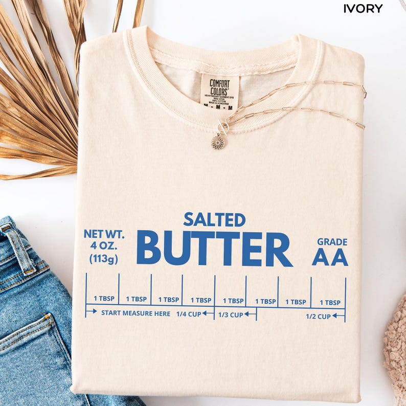 Op de afbeelding: Ivoor kleurig t-shirt met het woord "BUTTER" in grote blauwe letters. Het shirt heeft ook de tekst "SALTED", "NET WT. 113g", en "GRADE AA". Meetgidsen zijn onder het woord "BUTTER" gedrukt. Een gouden ketting is over het shirt gedrapeerd.