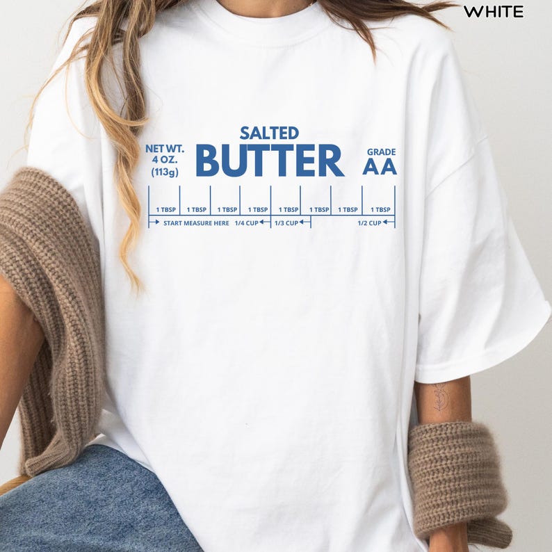 Op de afbeelding: Wit t-shirt met een blauwe opdruk met de tekst "SALTED BUTTER AA" en meetmarkeringen. Het shirt toont ook het nettogewicht van 113g.