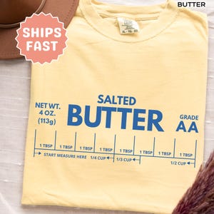 May include: A light yellow t-shirt with the word "BUTTER" in large blue letters. The shirt also features the text "SALTED", "GRADE AA", and a measurement guide. The shirt's net weight is listed as 4 oz (113g).