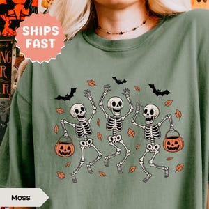 Op de afbeelding: Mosgroen T-shirt met een Halloween-ontwerp. De afbeelding toont drie dansende skeletten met pompoenemmers, met vleermuizen en herfstbladeren. Het shirt heeft de tekst "Moss" en een "Ships Fast" sticker.