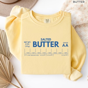 Op de afbeelding: Een lichtgeel sweatshirt met het woord "BUTTER" bovenaan. De voorkant van de trui heeft de woorden "SALTED BUTTER" in blauw, samen met meetmarkeringen. Het nettogewicht is 113 gram.