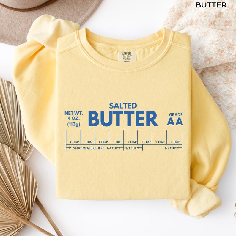 Op de afbeelding: Een lichtgeel sweatshirt met het woord "BUTTER" bovenaan. De voorkant van de trui heeft de woorden "SALTED BUTTER" in blauw, samen met meetmarkeringen. Het nettogewicht is 113 gram.