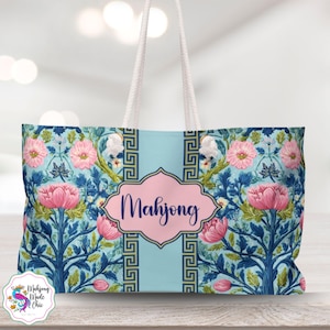 Könnte beinhalten: Eine große Tragetasche mit einem floralen Design in Blau- und Rosatönen. Die Tasche hat ein hellblaues Mittelteil mit dem Wort "Mahjong" in einem rosa, dekorativen Rahmen. Weiße Seilgriffe vervollständigen das Design.