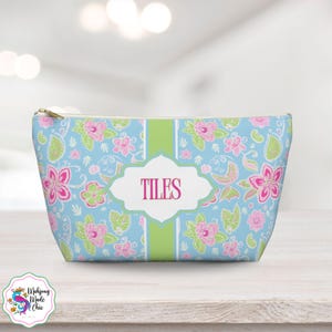 May include: A light blue cosmetic bag with a floral pattern in pink and green. The bag has a gold zipper and a white label with the name "TILES" in pink. The bag is decorated with a green stripe down the center.