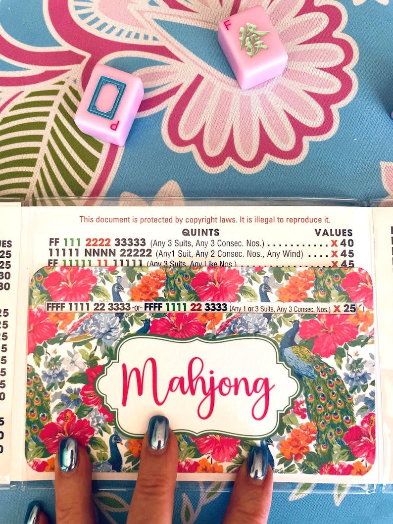 Pu&ograve; includere: Primo piano di un set di gioco del Mahjong. Il set include tessere rosa e una carta con la parola "Mahjong" in corsivo. Lo sfondo presenta un disegno floreale con tonalit&agrave; rosa e blu.