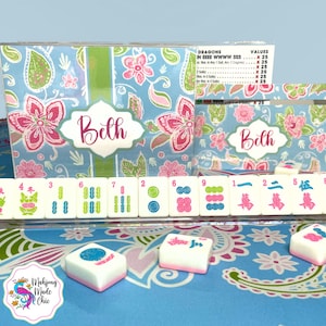 May include: A Mahjong set with a blue floral design, featuring pink flowers and paisley patterns. The set includes tiles and a box personalized with the name "Beth". Some tiles are visible, along with the text "Mahjong Made Chic".