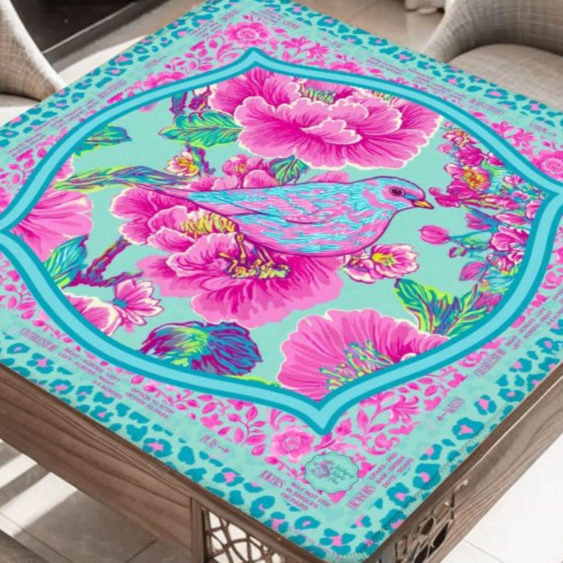 May include: A square decorative table cover with a vibrant floral and bird design. The cover features a teal background with a central framed design of a pink and blue bird surrounded by large pink flowers. The border has a leopard print pattern.