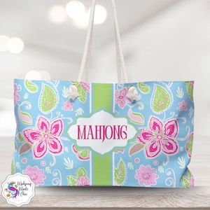 May include: A light blue tote bag with a floral pattern in pink and green. The bag features white rope handles and the word "MAHJONG" in a white and pink label. The bag has a green stripe down the center.