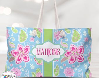 May include: A light blue tote bag with a floral pattern in pink and green. The bag features white rope handles and the word "MAHJONG" in a white and pink label. The bag has a green stripe down the center.