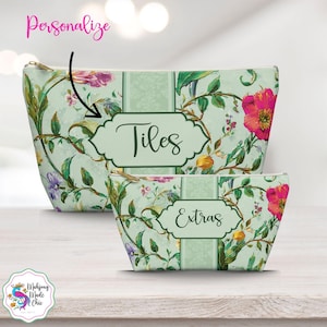 May include: Two floral print cosmetic bags. The larger bag has the word "Tiles" in a decorative label, and the smaller bag has the word "Extras". The bags have a light green background with pink, purple, and yellow flowers.