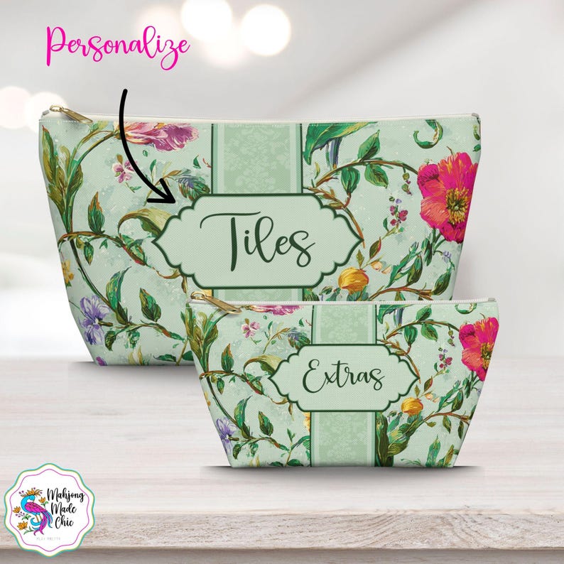 K&ouml;nnte beinhalten: Zwei Kosmetiktaschen mit Blumendruck. Die gr&ouml;&szlig;ere Tasche tr&auml;gt das Wort &bdquo;Tiles&ldquo; in einem dekorativen Etikett, und die kleinere Tasche tr&auml;gt das Wort &bdquo;Extras&ldquo;. Die Taschen haben einen hellgr&uuml;nen Hintergrund mit rosa, lila und gelben Blumen.