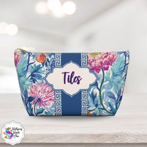 May include: A floral print cosmetic bag with a gold zipper. The bag features a blue and white background with pink and purple flowers and the word "Tiles" in a white and blue frame. The bag is on a wooden surface.