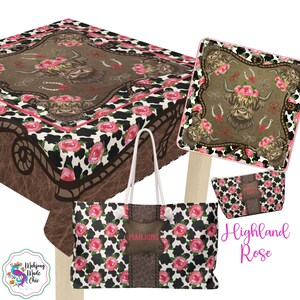 May include: A collection of Highland Rose themed items, including a tablecloth, tote bag, and cosmetic bag. The items feature a cowhide pattern with pink roses and a brown background with a Highland cow illustration. The tote bag has a brown leather-like patch with the name "Marjone".