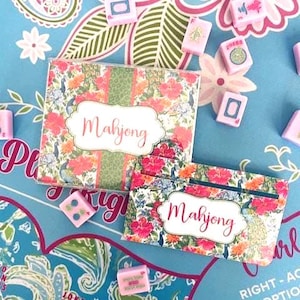 Pu&ograve; includere: Un set di Mahjong con disegni floreali e piume di pavone. Il set include una scatola e una custodia, entrambe con la scritta "Mahjong". Tessere di Mahjong rosa e bianche sono sparse su uno sfondo blu.