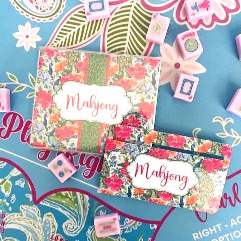Pu&ograve; includere: Un set di Mahjong con disegni floreali e piume di pavone. Il set include una scatola e una custodia, entrambe con la scritta "Mahjong". Tessere di Mahjong rosa e bianche sono sparse su uno sfondo blu.