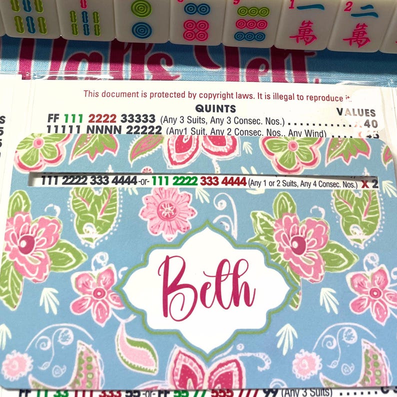 Pu&ograve; includere: Un porta carte Mahjong personalizzato con un design floreale rosa, verde e bianco su sfondo azzurro. Il nome "Beth" &egrave; visualizzato in corsivo. Il porta carte &egrave; posizionato vicino alle tessere del Mahjong.