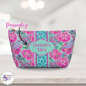Op de afbeelding: Een gepersonaliseerde make-uptas met een bloemen- en vogelpatroon. De tas heeft een turquoise achtergrond met roze bloemen en blauwe vogels. Het midden heeft een turquoise paneel met de tekst "Hannah's Tiles".