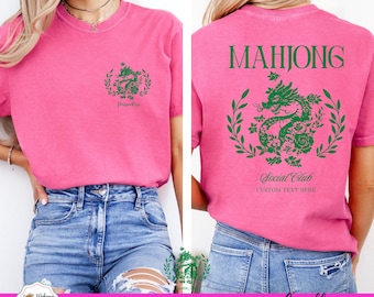 May include: A pink t-shirt with a green dragon design and the word "Mahjong" on the back. The front of the shirt has a smaller dragon design. The shirt also has the words "Social Club" and "Custom Text Here".