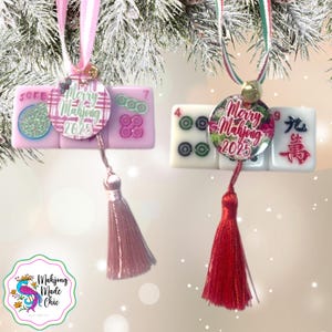 May include: Two Mahjong tile ornaments with tassels. One is pink with a pink and white ribbon, the other is white with a green and red ribbon. Each has a circular charm that says "Merry Mahjong 2025".