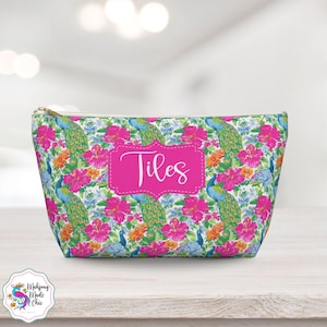 Può includere: Un beauty case con un vivace motivo floreale e di piume di pavone. La borsa presenta un rettangolo rosa con il nome "Tiles" in corsivo bianco. Il design include fiori di ibisco rosa, fiori d'arancio e pavoni blu su sfondo bianco.