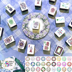 May include: A collection of white Mahjong tiles with various symbols and numbers, arranged around a decorative floral plate. The tiles are scattered on a blue and white checkered surface. A logo with the text "Mahjong Made Chic" is visible.
