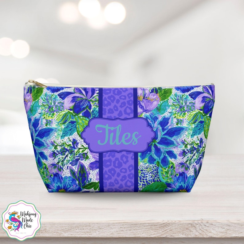 May include: A zippered cosmetic bag with a floral pattern in shades of blue, purple, and green. The bag features a purple stripe with a leopard print and the name "Tiles" in a light blue script. The bag has a gold zipper.