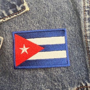 May include: A blue denim jacket featuring a rectangular embroidered patch of the Cuban flag. The patch displays a red triangle on the left side with a white star, followed by three blue horizontal stripes and two white horizontal stripes.