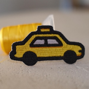 May include: Embroidered yellow taxi cab patch with black trim and wheels. The patch has a small roof light and two windows. A spool of yellow thread is in the background.