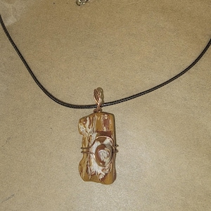 Può includere: Collana con pendente fatto a mano con una pietra rettangolare marrone chiaro con un motivo unico. La pietra è avvolta con filo di rame e appesa a una collana di cordino nero con chiusura argentata.