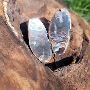 May include: Two polished agate stones with a natural, layered pattern of white, brown, and gray. The stones are oval-shaped and have a hole drilled at the top for jewelry making. They rest on a piece of weathered wood.