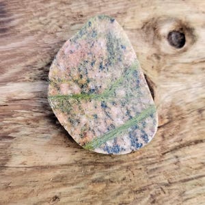 Könnte beinhalten: Ein polierter, tropfenförmiger Stein mit einem gesprenkelten rosa, grünen und schwarzen Muster. Der Stein hat grüne Adern, die über seine Oberfläche verlaufen. Der Stein wird auf einer verwitterten Holzoberfläche gezeigt.