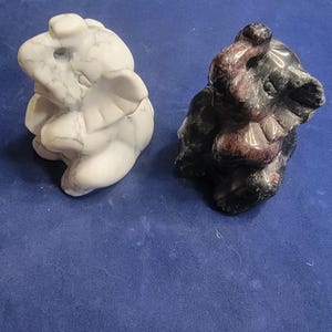 May include: Two stone sculptures of abstract figures. One is white with gray veining, the other is black with red marbling. Both are on a blue surface. The sculptures have a rounded, organic shape.