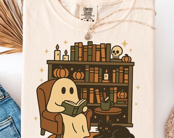 May include: Cream-colored t-shirt featuring a cartoon ghost reading a book in a cozy setting. The design includes a bookshelf, pumpkins, candles, a black cat, and a coffee mug. The shirt has a Comfort Colors tag.