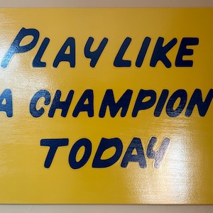 May include: Yellow wooden sign with the text "PLAY LIKE A CHAMPION TODAY" in dark blue lettering. The sign has a glossy finish and is mounted with small, dark-colored screws in each corner.